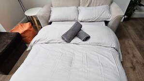 Een opgemaakt bed met een opgerolde handdoek, een klein oranje kussen en een donkere bank.