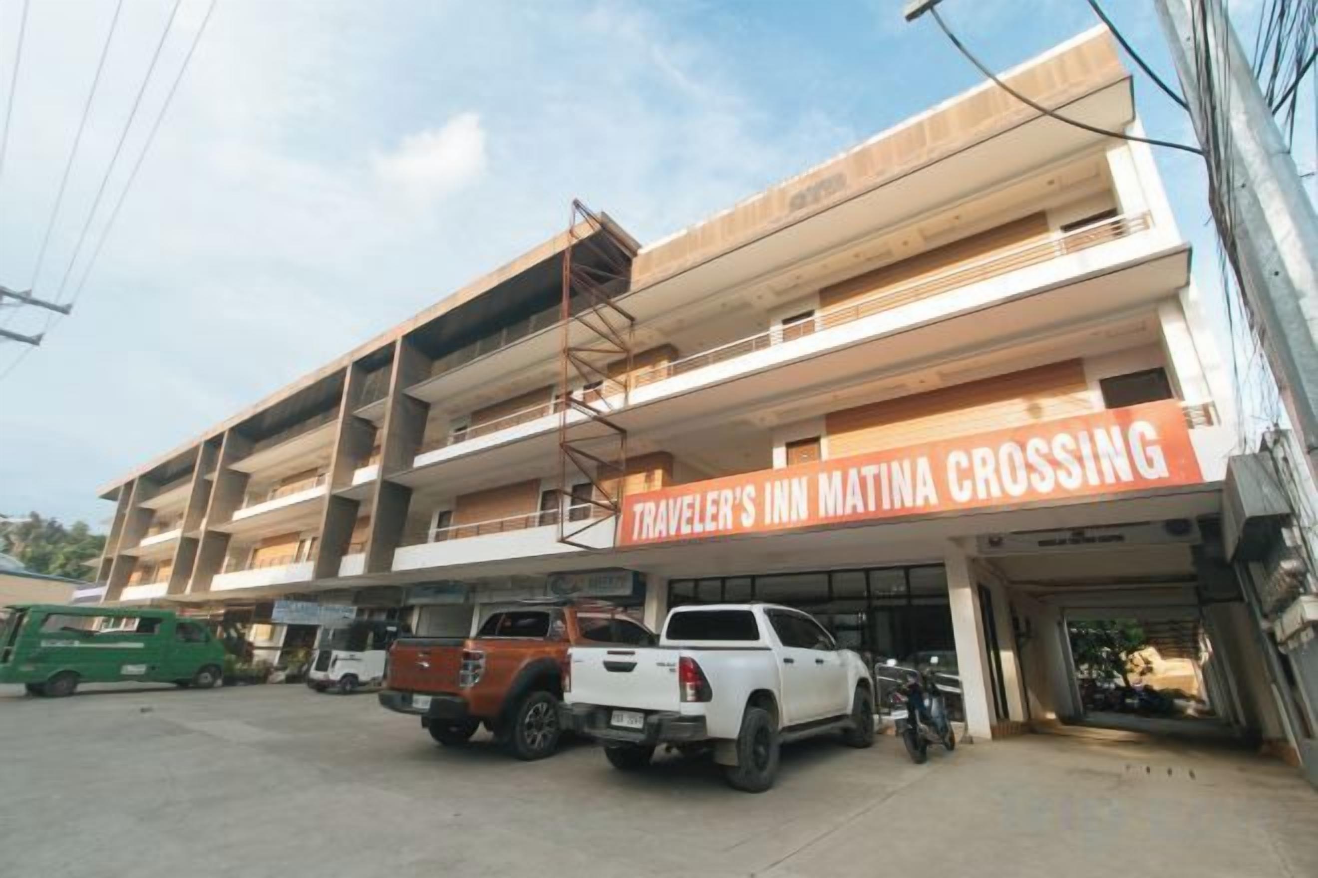 A multi-story building with a sign for 'Traveler's Inn Matina Crossing.' There are vehicles parked in front, and the building has multiple balconies.