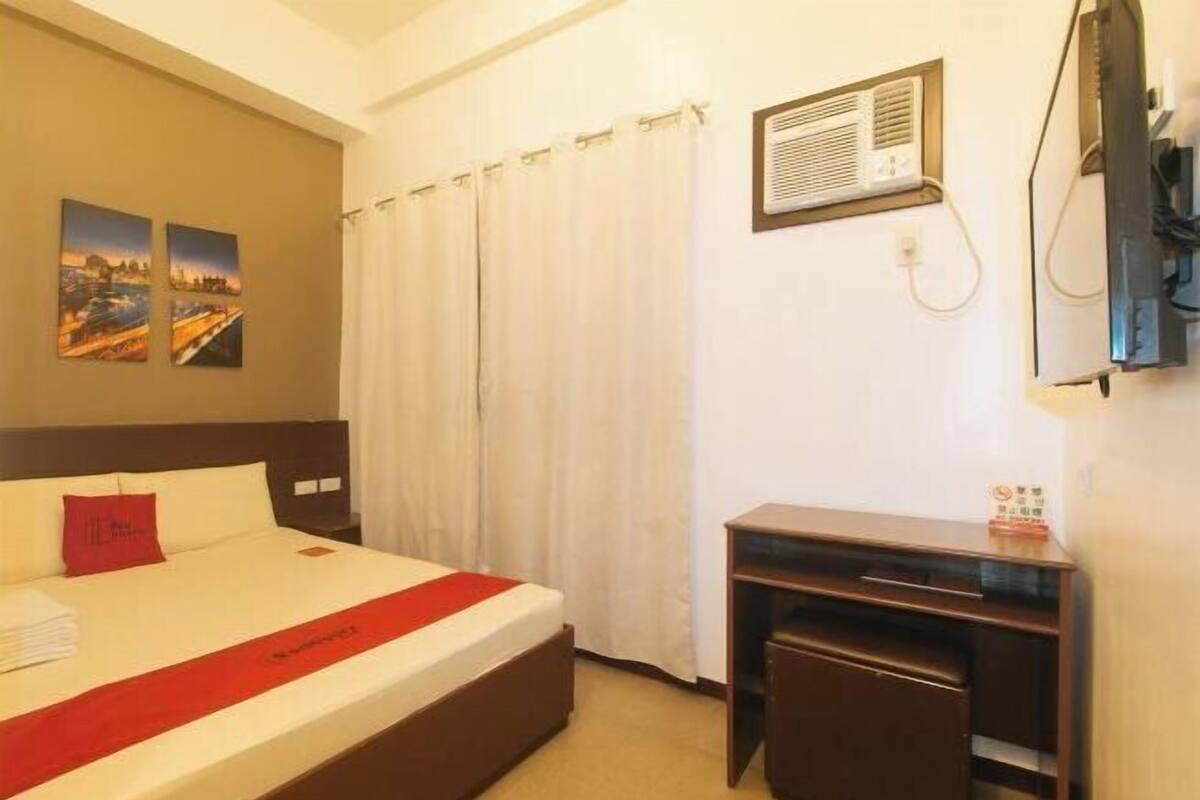 A hotel room with a bed, a desk, a wall-mounted air conditioner, and two paintings on the wall.
