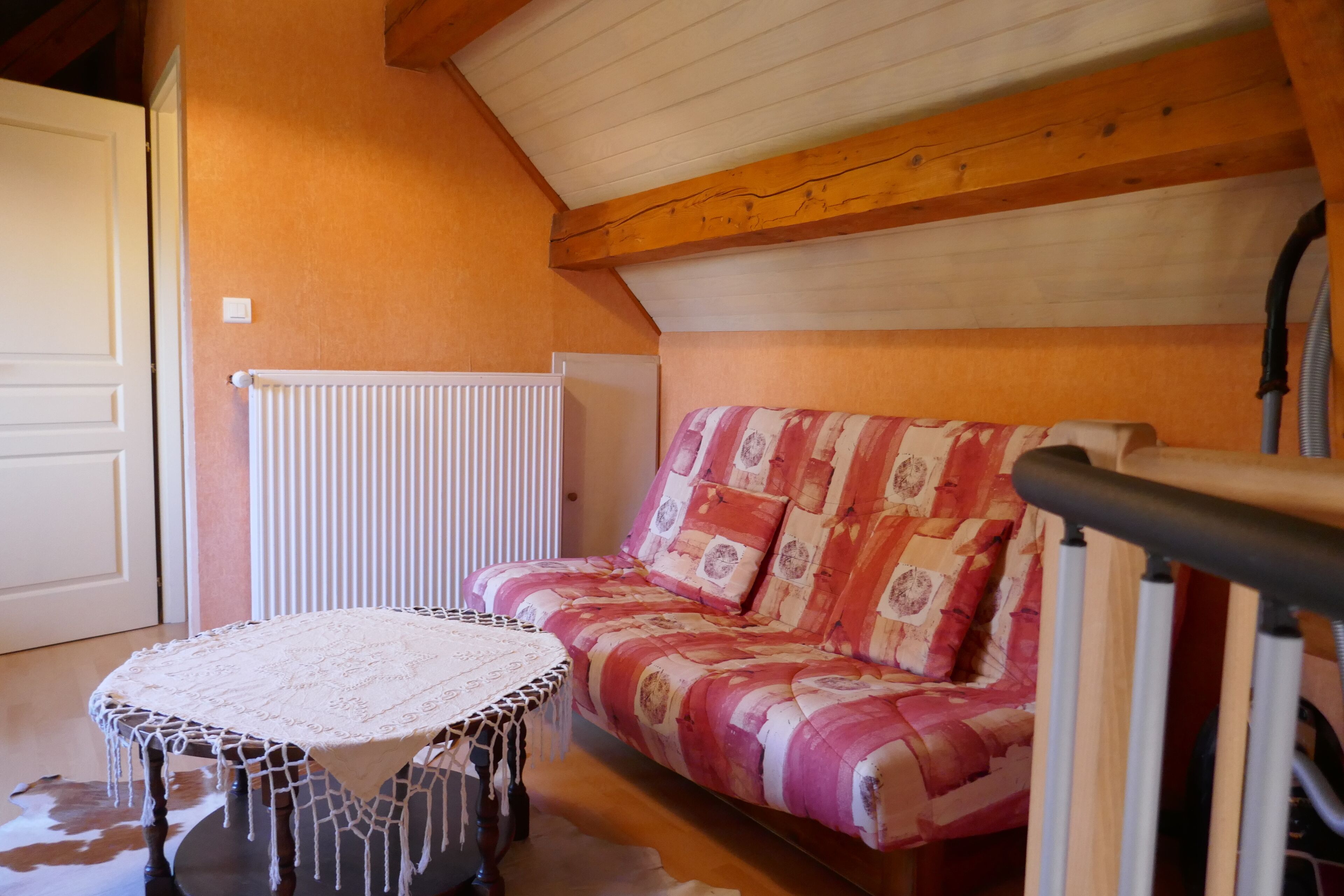 Ein gemütliches Zimmer mit Holzbalkendecke, einem gemusterten Sofa, einem kleinen runden Tisch mit Spitze und einem Heizkörper.