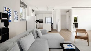 A modern living room with a grey sofa, a wooden chair, and a coffee table. There is a kitchen in the background with white cabinets and a refrigerator. - Penthouse - Four Bedroom Apartment, Sleeps 19 (Jerusalem)