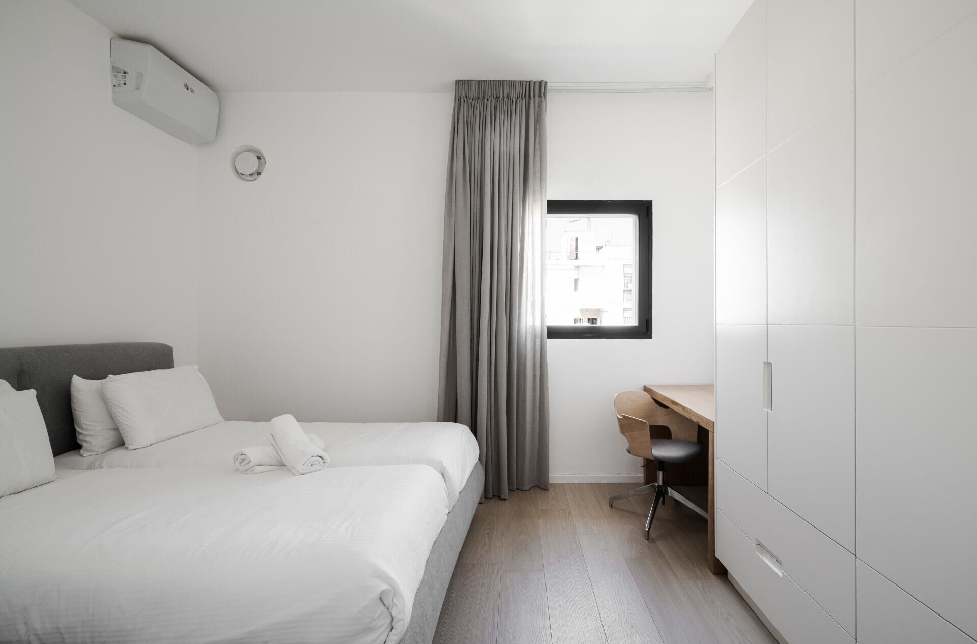 A hotel room with a bed, a desk, a wardrobe, and a window with a view of buildings.