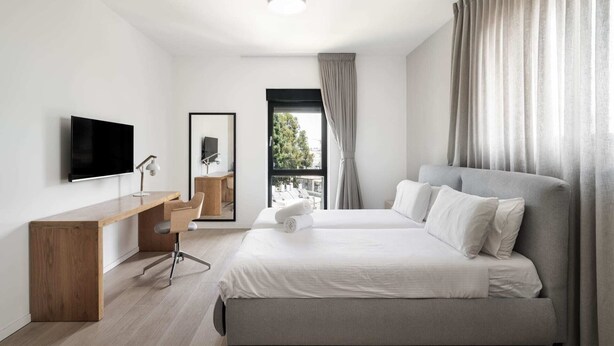A modern hotel room with a large bed, a desk with a chair, a flat-screen TV, and a window with curtains. - Penthouse - Four Bedroom Apartment, Sleeps 19 (Jerusalem)