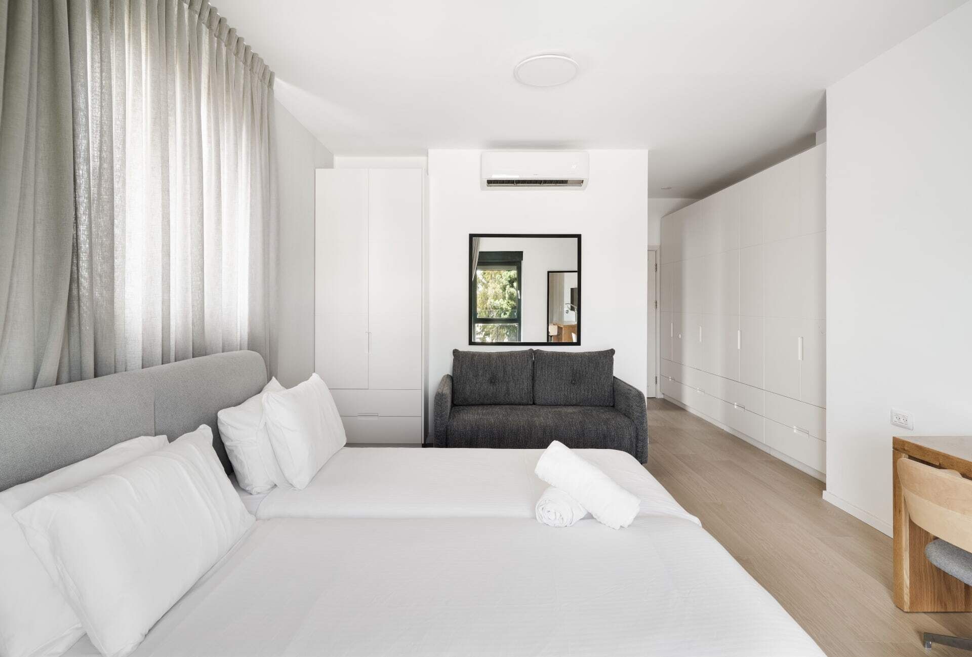 Ein modernes Hotelzimmer mit einem großen Bett, einem grauen Sofa, einem Kleiderschrank und einem Spiegel, der einen Balkon widerspiegelt.