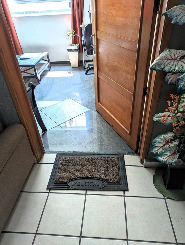 A tiled entrance with a welcome mat, a wooden door, a small table with a chair, and a potted plant.