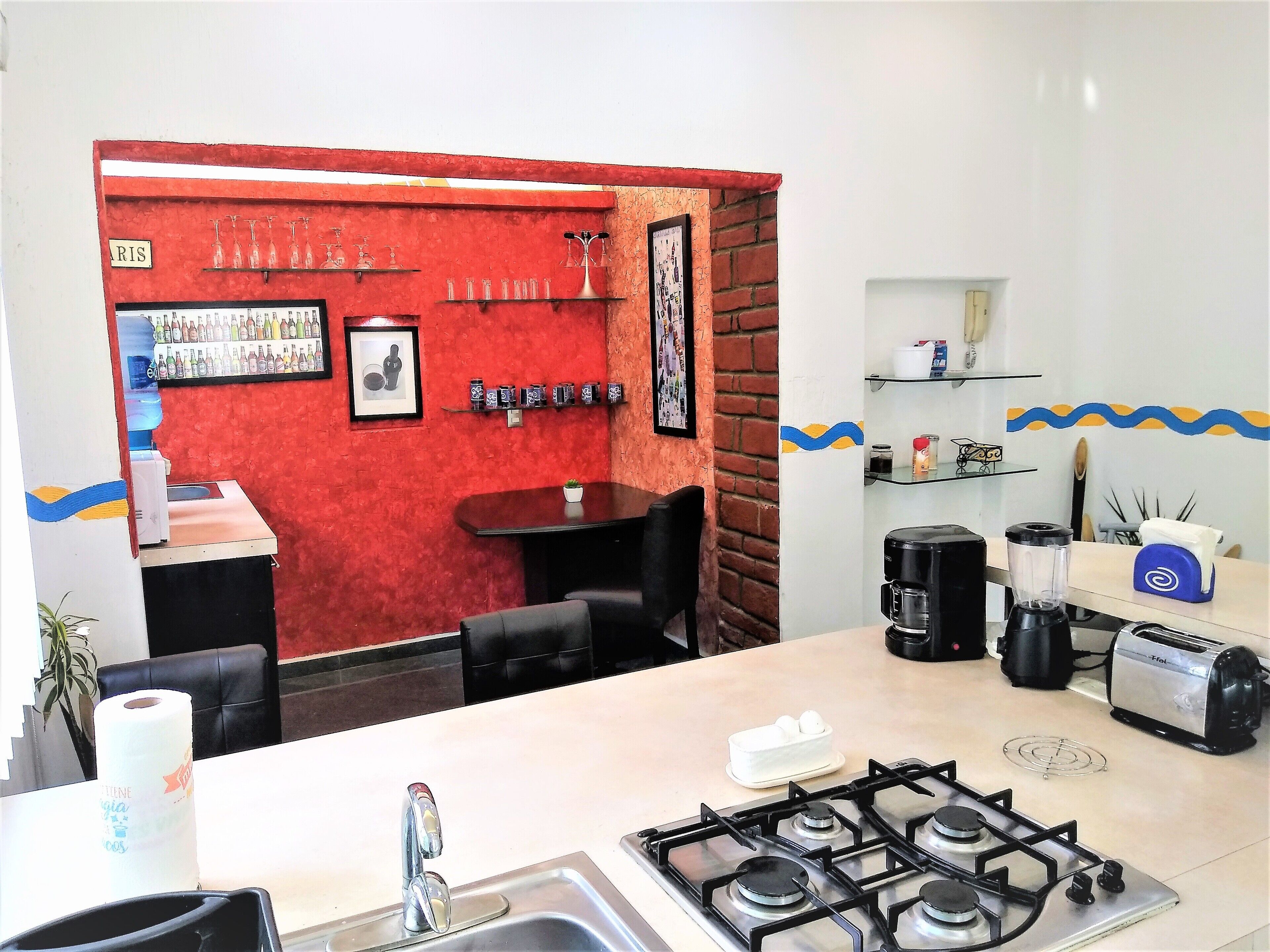 A kitchen with a gas stove, a sink, and a countertop with a toaster. There is a red brick wall with shelves holding glasses and a framed picture.