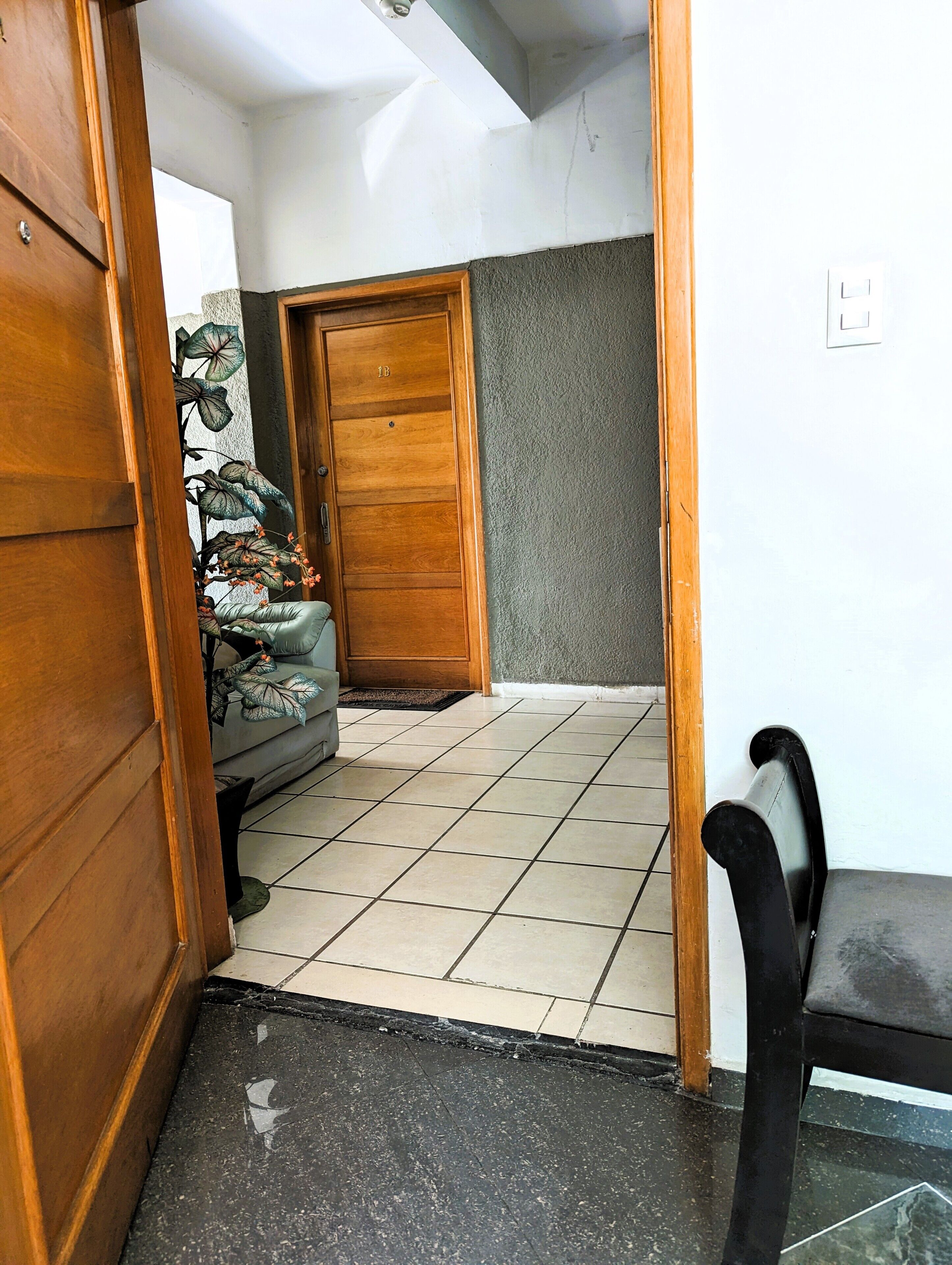 A room with a wooden door, a tiled floor, a grey sofa, and a black chair.