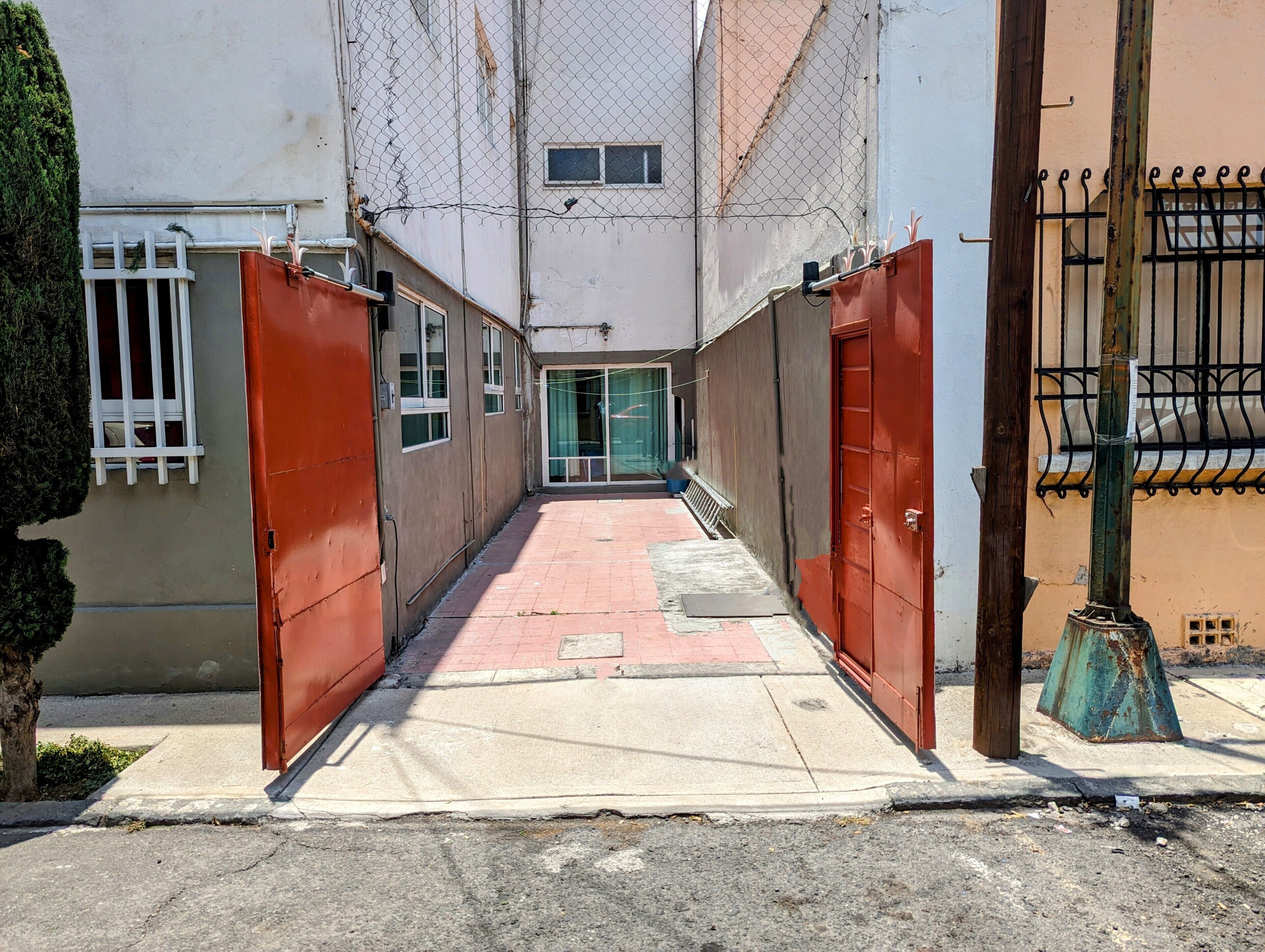 A narrow alleyway with closed red gates, a concrete pathway, and a building with a window and a door.