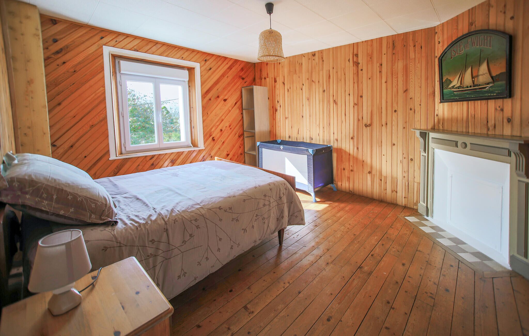 Een slaapkamer met houten wanden, een bed, een nachtkastje, een lamp, een dressoir, een raam en een schilderij van een zeilboot.
