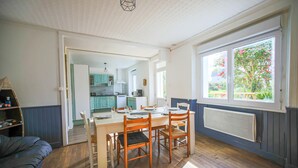A dining area with a wooden table and chairs, a kitchen in the background, and a window with a view of greenery. - 3 bedroom stunning home in Plougasnou (Plougasnou)