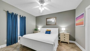 A bedroom with a bed, a nightstand, a ceiling fan, a window with curtains, and a wall-mounted mirror. - Twin Palms 603 (Panama City Beach)