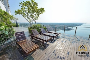 Een dak terras met houten lounge fauteuils en een glazen balustrade met uitzicht op een stadsbeeld.