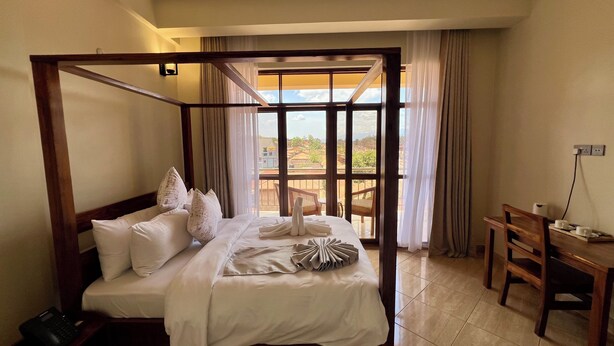 Una cama con dosel cubierta con sábanas y almohadas blancas, un escritorio de madera con una silla, y vistas de un paisaje urbano a través de amplios ventanales.