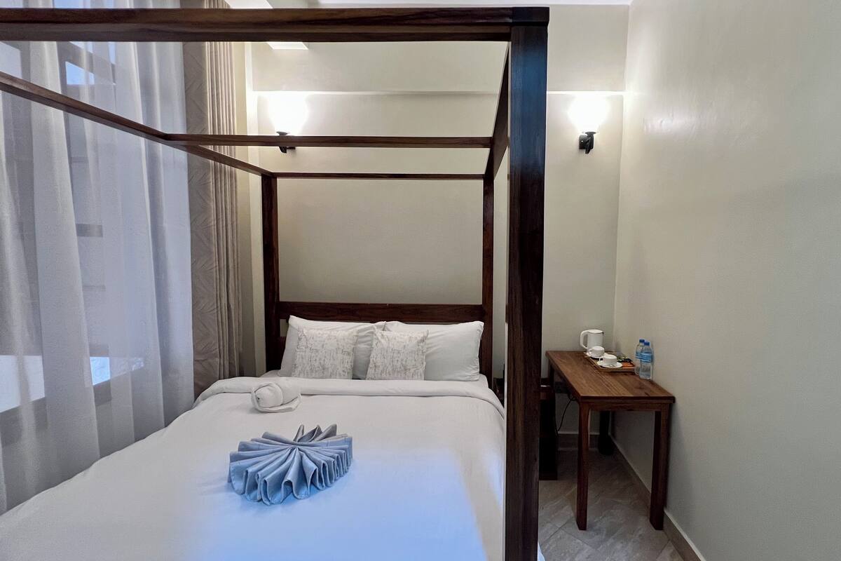 Ein Himmelbett mit weißer Bettwäsche, ein Nachttisch mit Tasse und Wasserflasche sowie eine Wandleuchte.