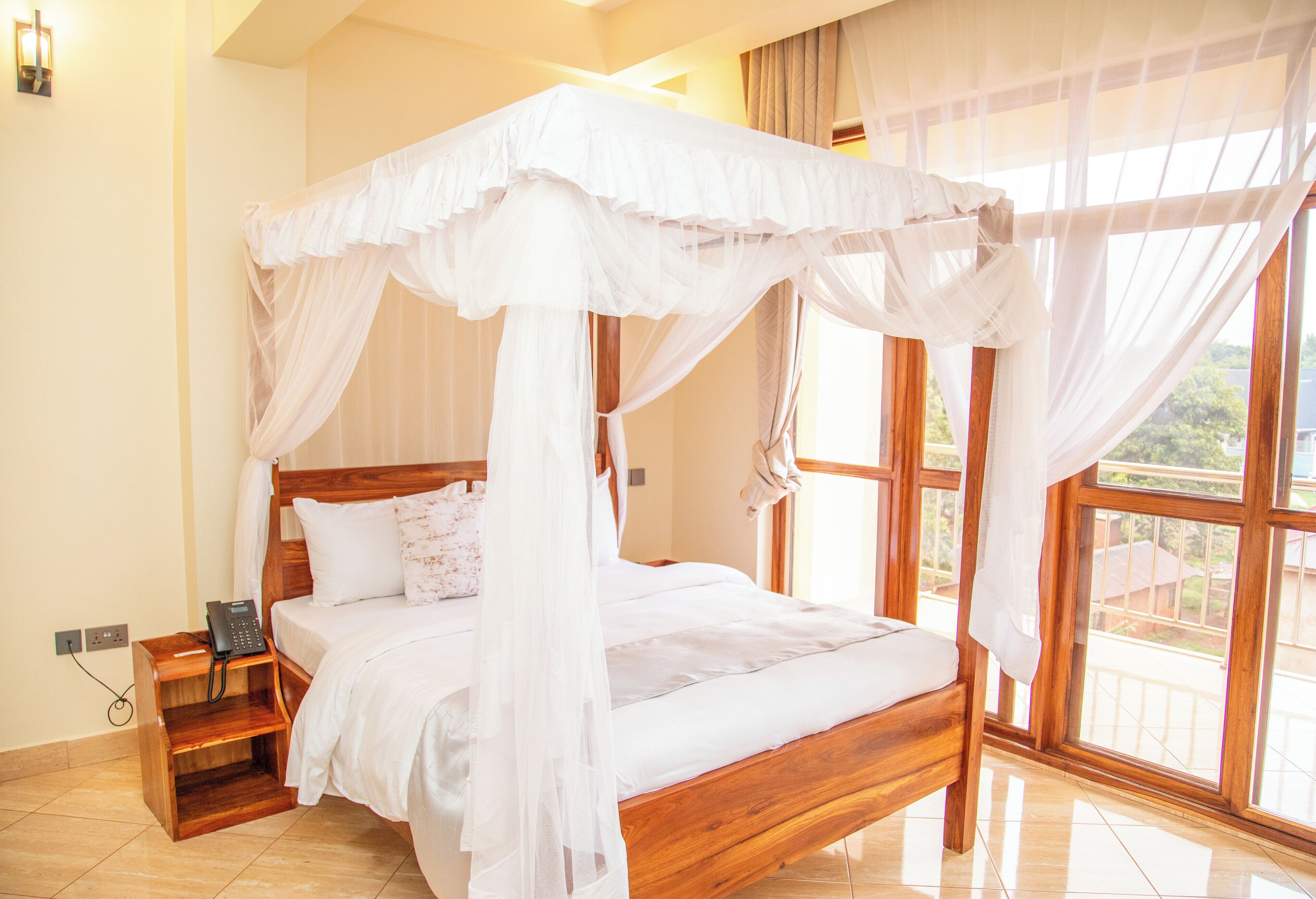 A four-poster bed with a canopy, a bedside table with a phone, and a view of the outdoors through large windows.