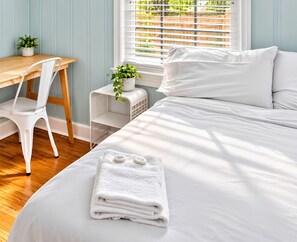 Una cama bien tendida con sábanas blancas y una toalla doblada, una silla blanca, una mesita con una planta y una ventana con persianas.
