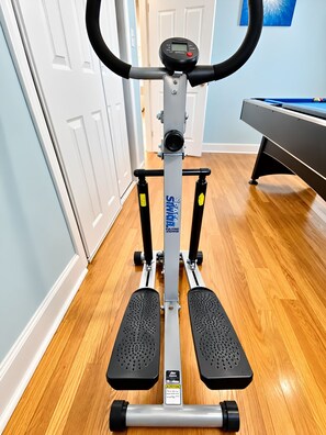 A stationary bike with a digital display, positioned in a room with wooden flooring, a pool table, and a door in the background. - Central Wilmington Retreat: Pet-Friendly, Family Comfort & Loved by Guests (Wilmington)