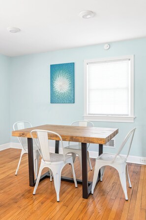 Un comedor con mesa de madera y sillas blancas, pared azul y una ventana con persianas.