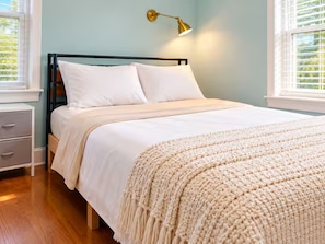 Una cama con ropa de cama blanca, un cabecero de madera y una mesita de noche blanca con cajones.