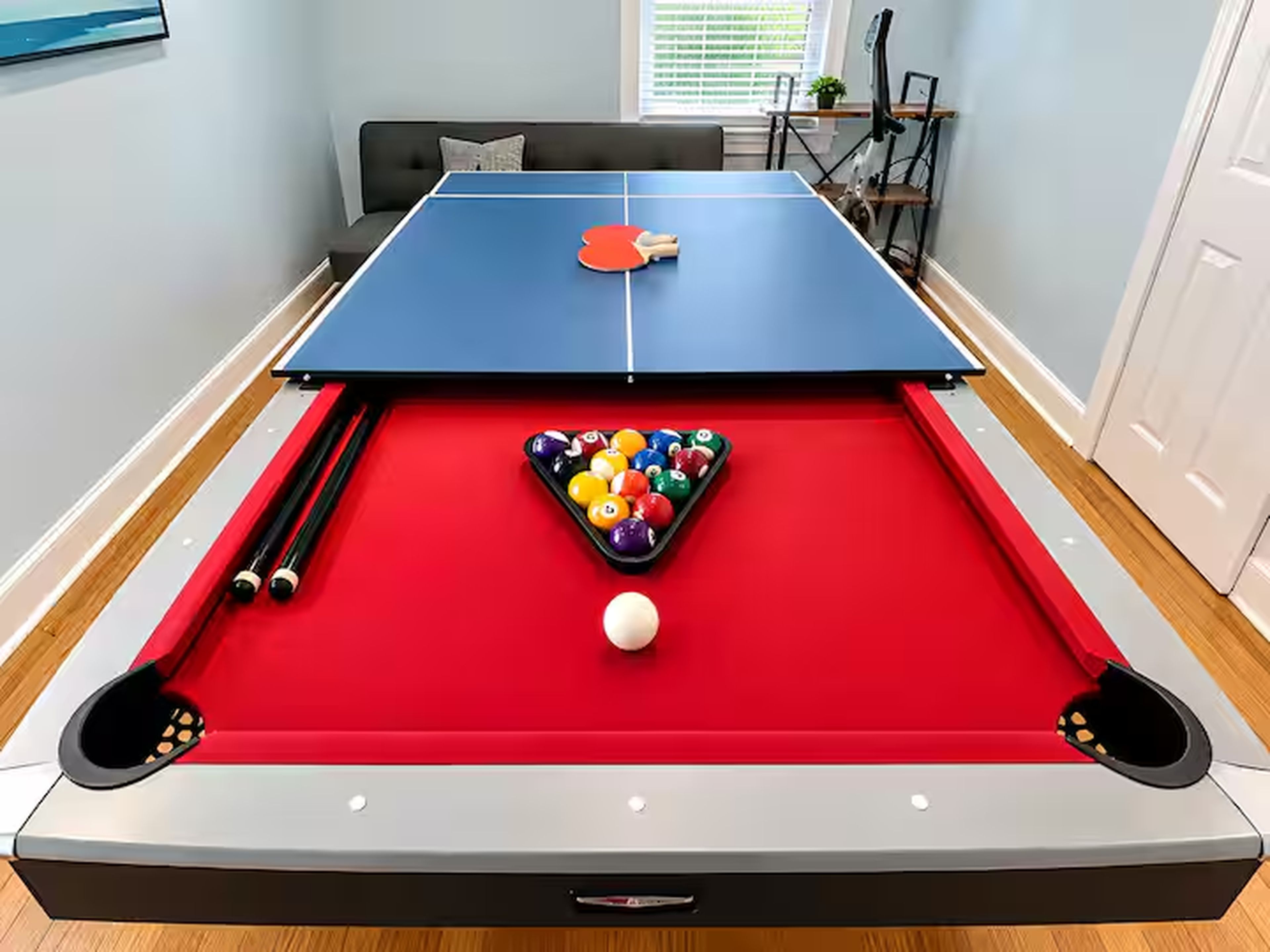 Een pooltafel met een rode viltkleed, omgeven door een grijze rand, en een set poolkeus.