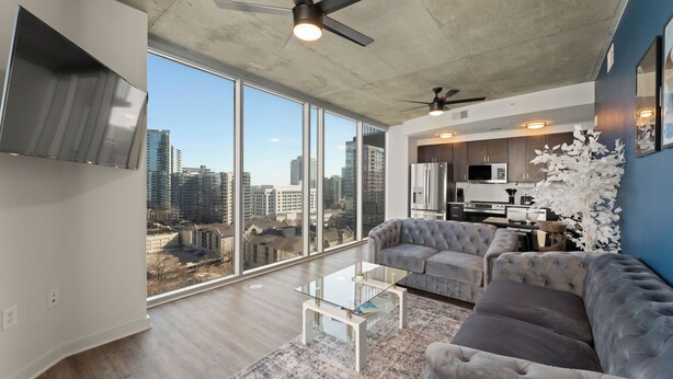 A spacious living room with a large window, a grey sofa, a glass coffee table, and a ceiling fan. - 19th Floor Midtown Sky Suite | Free Parking | Gym (Atlanta)