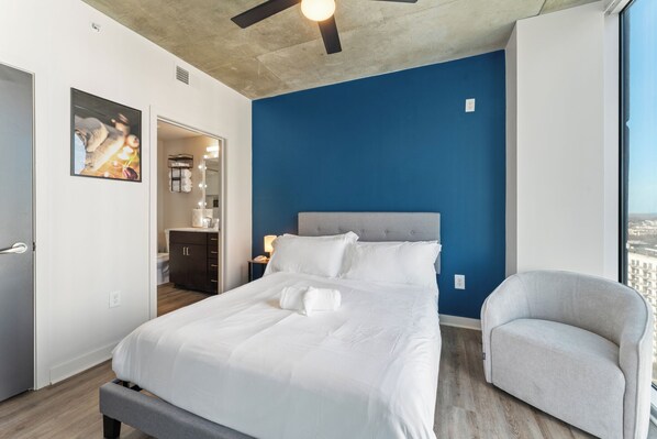 A modern hotel room with a large bed, a grey armchair, and a view of the city through a large window.