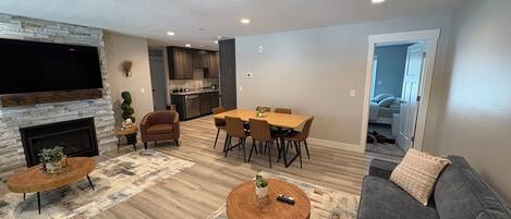 A living room with a fireplace, a TV mounted on the wall, a dining area with a table and chairs, and a gray sofa.