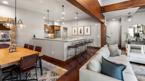 Un salón amplio con una viga de madera grande, un comedor con mesa y sillas de madera, y una cocina con electrodomésticos modernos e iluminación colgante.