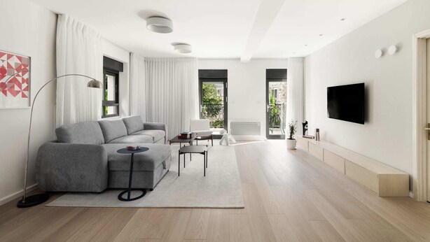A modern living room with a grey sectional sofa, a small round table, a floor lamp, and a flat-screen TV mounted on the wall.