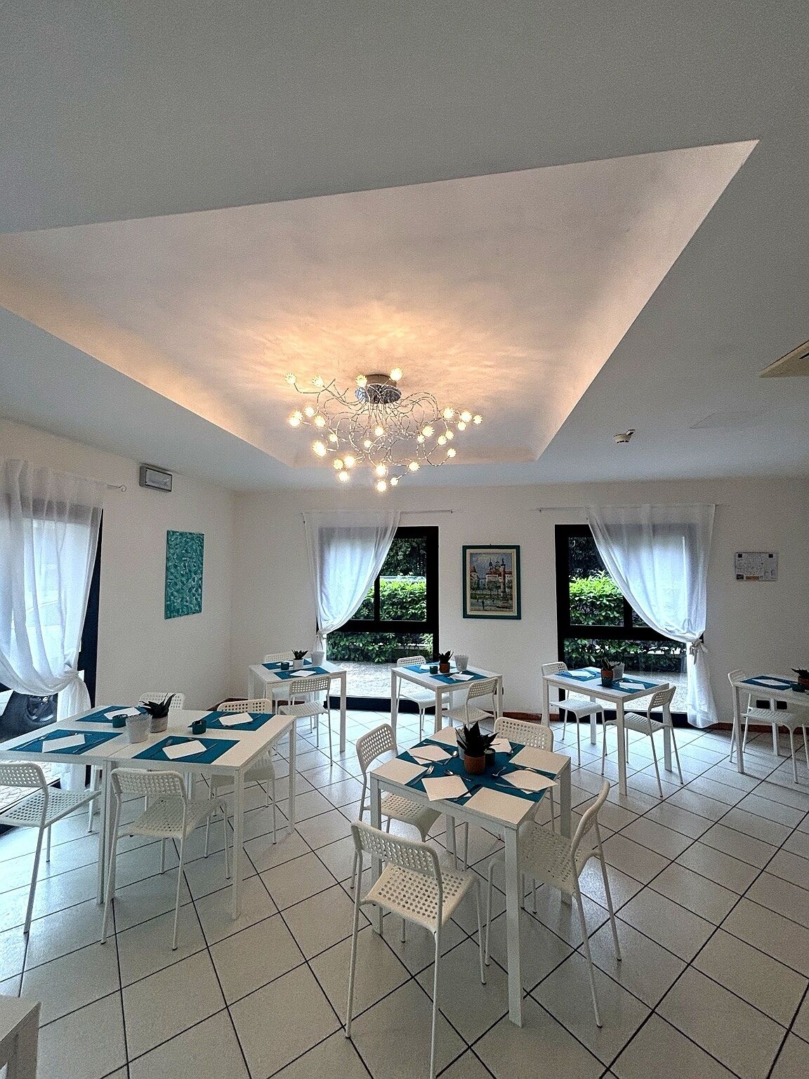 A dining area with white tables and chairs, a chandelier, and large windows with curtains.