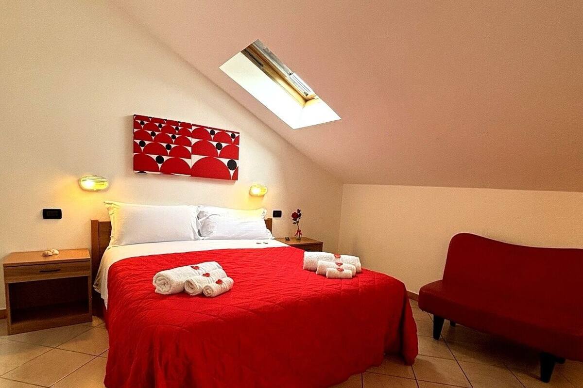 A hotel room with a bed, a red sofa, a wall-mounted artwork, a nightstand, and a ceiling light.