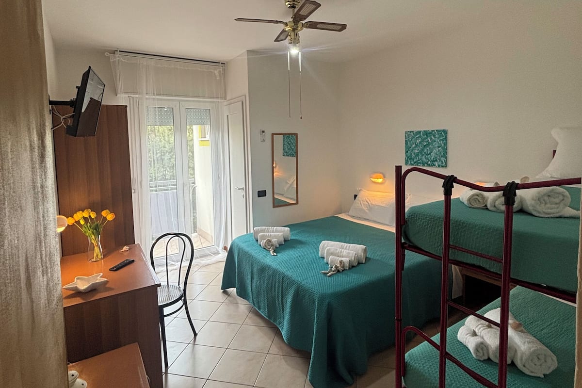 A room with a desk, a bed with a green bedspread, a bunk bed, a ceiling fan, and a window with curtains.