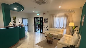 A reception area with a teal-colored counter, a white sofa, a coffee table, and a potted plant. - Hotel Dream (Rimini)
