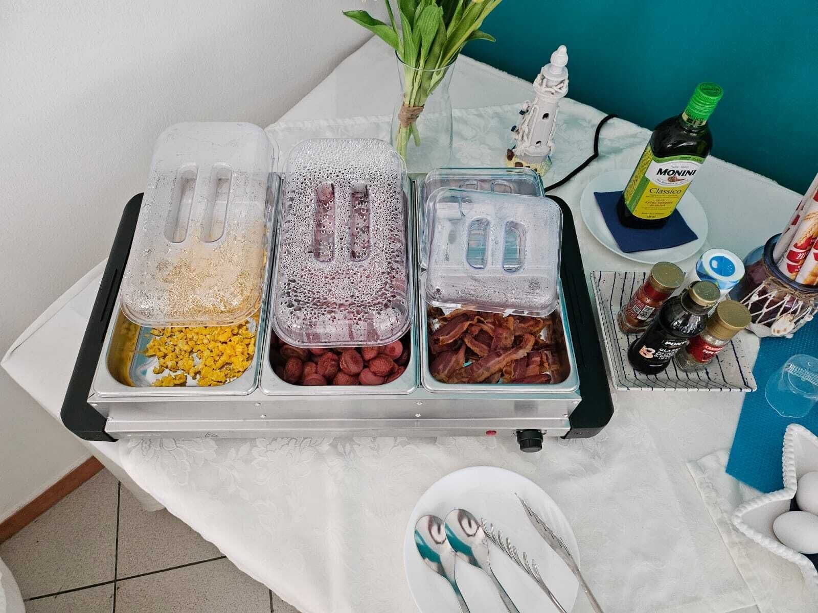 A buffet table with three containers of food, a bottle of Monini Classic, condiments, and utensils.