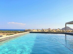A swimming pool with clear blue water, surrounded by a low wall and a view of a coastal area with buildings and mountains in the background.