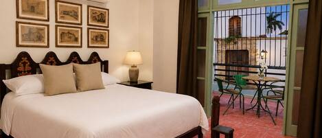 A hotel room with a bed, a nightstand, a lamp, a balcony with chairs, and framed pictures on the wall.