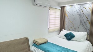 A bedroom with a bed, a sofa, a window with blinds, and an air conditioning unit on the wall.