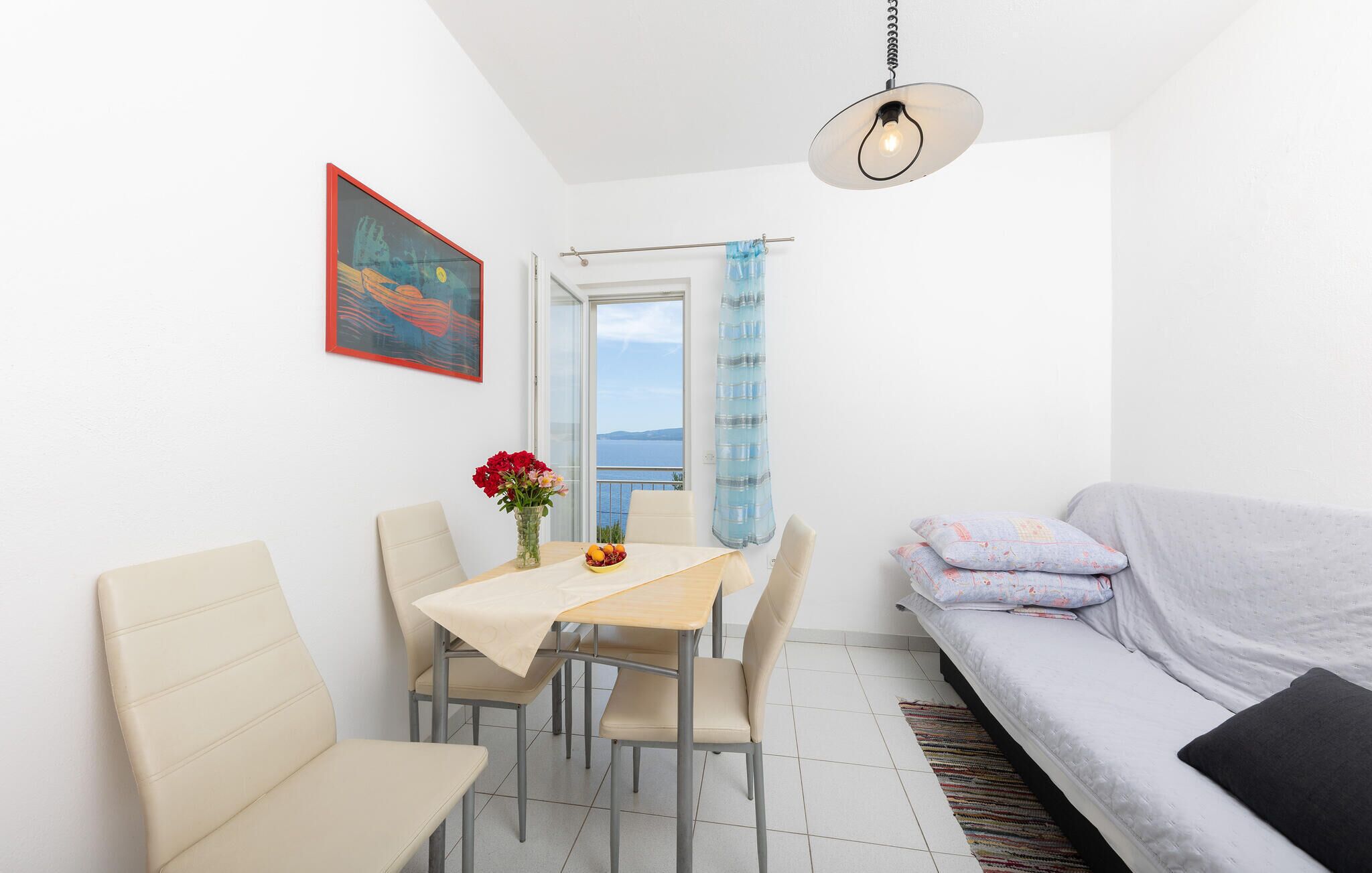 A small, bright room with a dining table, chairs, a sofa, and a view of the sea.