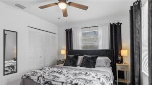Quarto com cama, ventilador de teto, janela com cortinas, mesa de cabeceira e espelho.