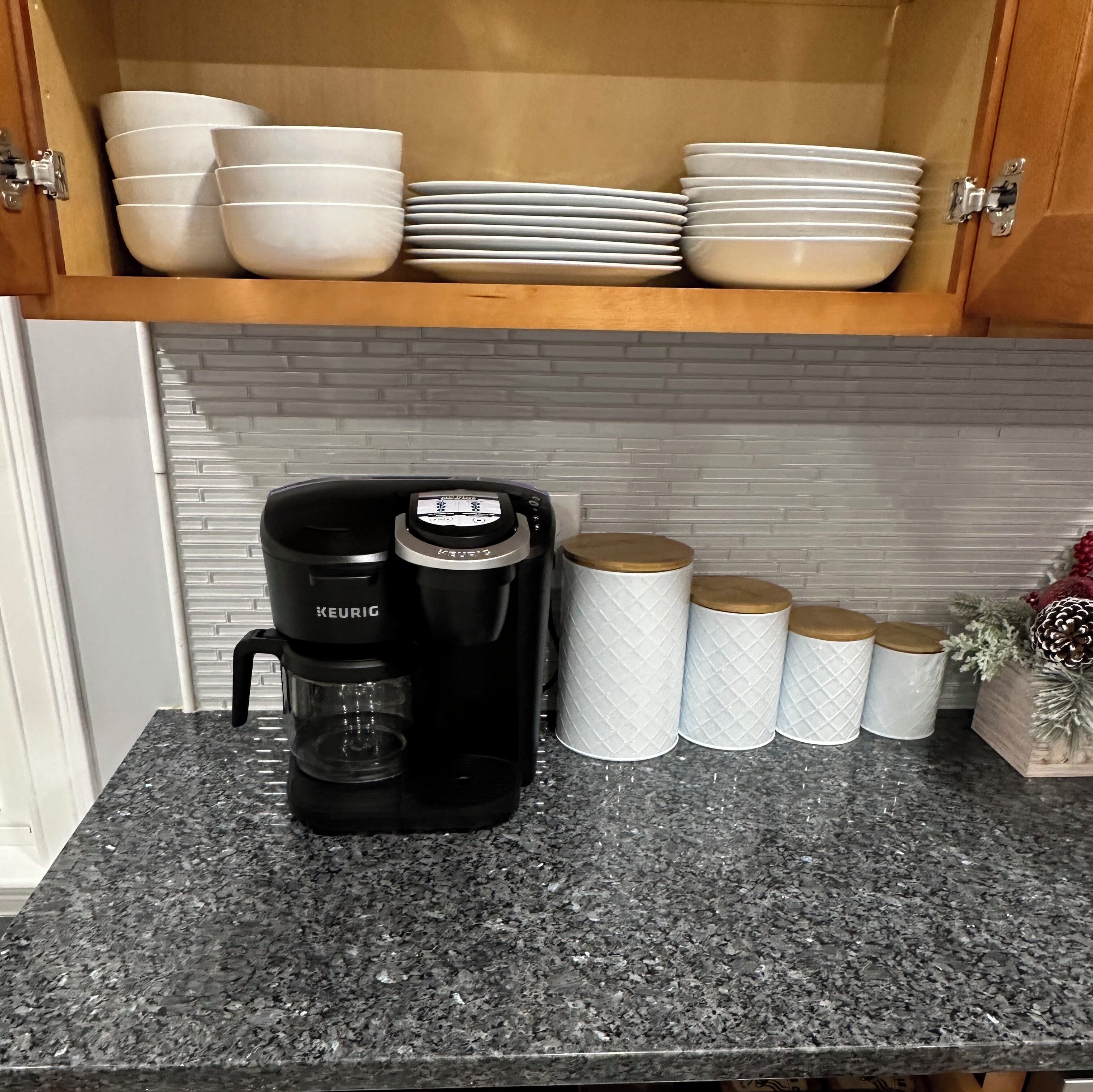 Une cuisine avec une machine à café Keurig noire posée sur un comptoir en granit, des bols et des assiettes blancs empilés au-dessus, et des boîtes de rangement blanches avec des couvercles en bois.