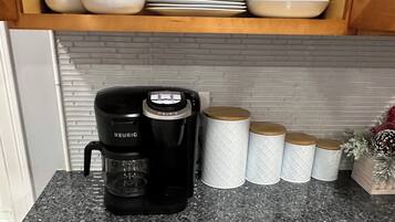 Une cuisine avec une machine à café Keurig noire posée sur un comptoir en granit, des bols et des assiettes blancs empilés au-dessus, et des boîtes de rangement blanches avec des couvercles en bois.