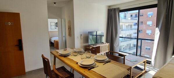 A dining area with a wooden table set for four, a television on a stand, and a large window with a view of a building.