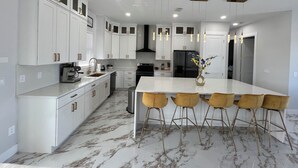 Una cocina moderna con armarios blancos, encimera de mármol, una isla central con taburetes y electrodomésticos de acero inoxidable.