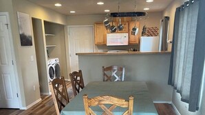 Zona pranzo con tavolo e sedie in legno, cucina con mobili in legno e rastrelliera pensile per pentole, e locale lavanderia con lavatrice.