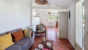 A spacious living room with a grey sofa, a chair, and a coffee table with cups and books. There is a large window and a door leading to another room. - Maison 5 Cantons Anglet (ANGLET)