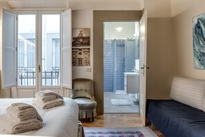 A bedroom with a bed, a chair, a bedside table, a nightstand, a window, and a door leading to a bathroom.
