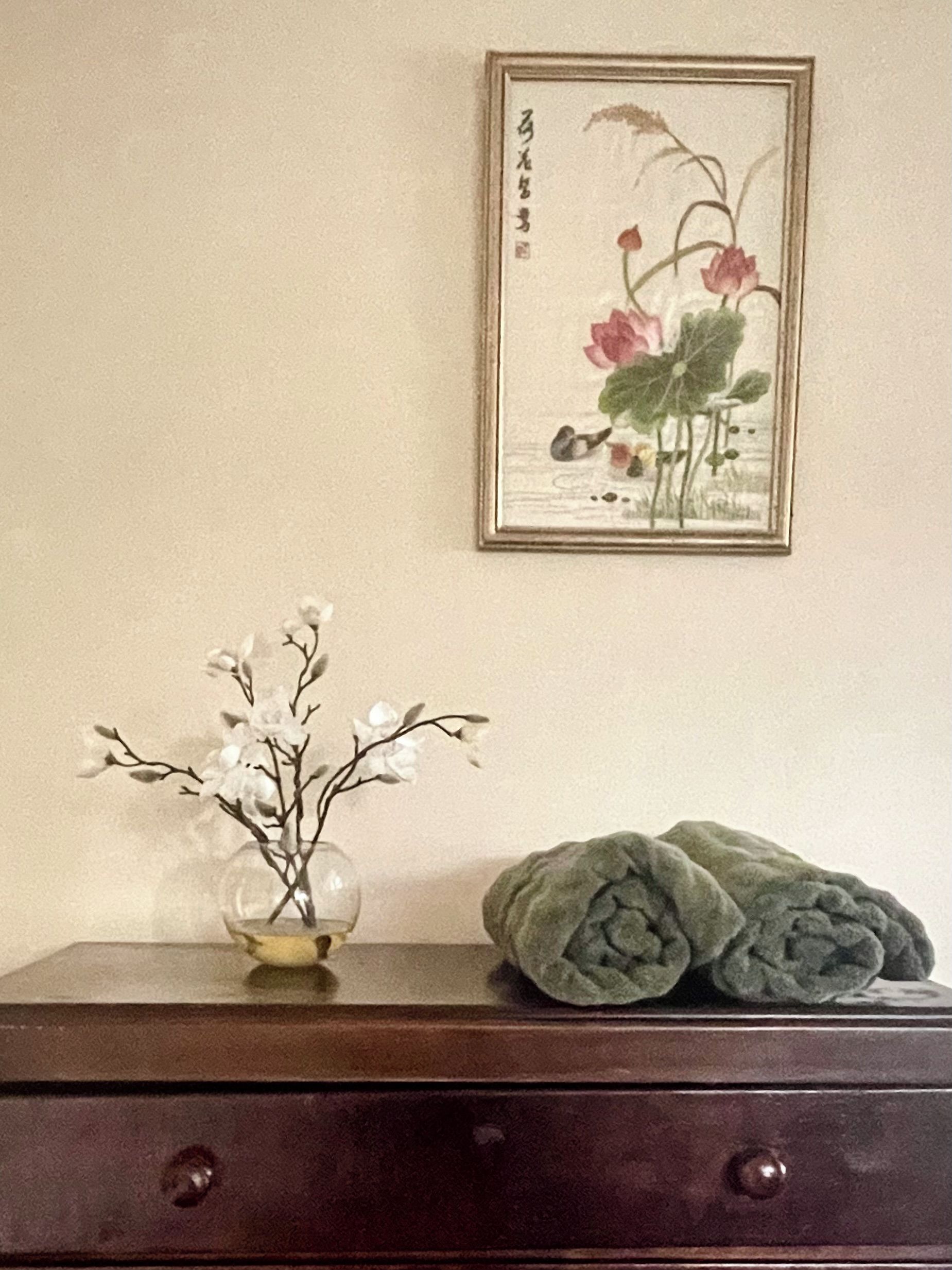 Une commode en bois, sur laquelle est posé un vase de fleurs blanches, et un tableau encadré représentant des fleurs roses, accroché au mur.