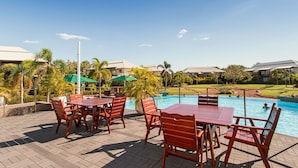 Ein Poolbereich mit Holztischen und -stühlen, ein Schwimmbecken und im Hintergrund der Blick auf einen Golfplatz und Gebäude.