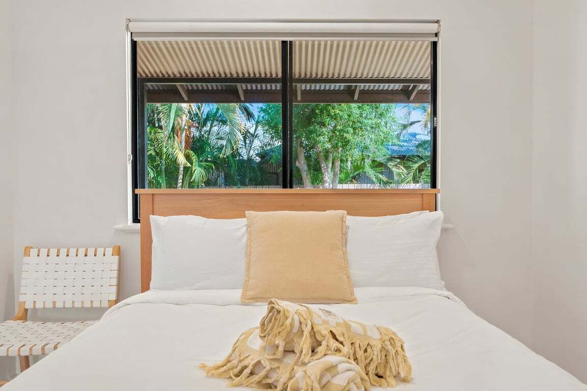 A bedroom with a bed, a window with a view of greenery, a wooden headboard, and a white chair.