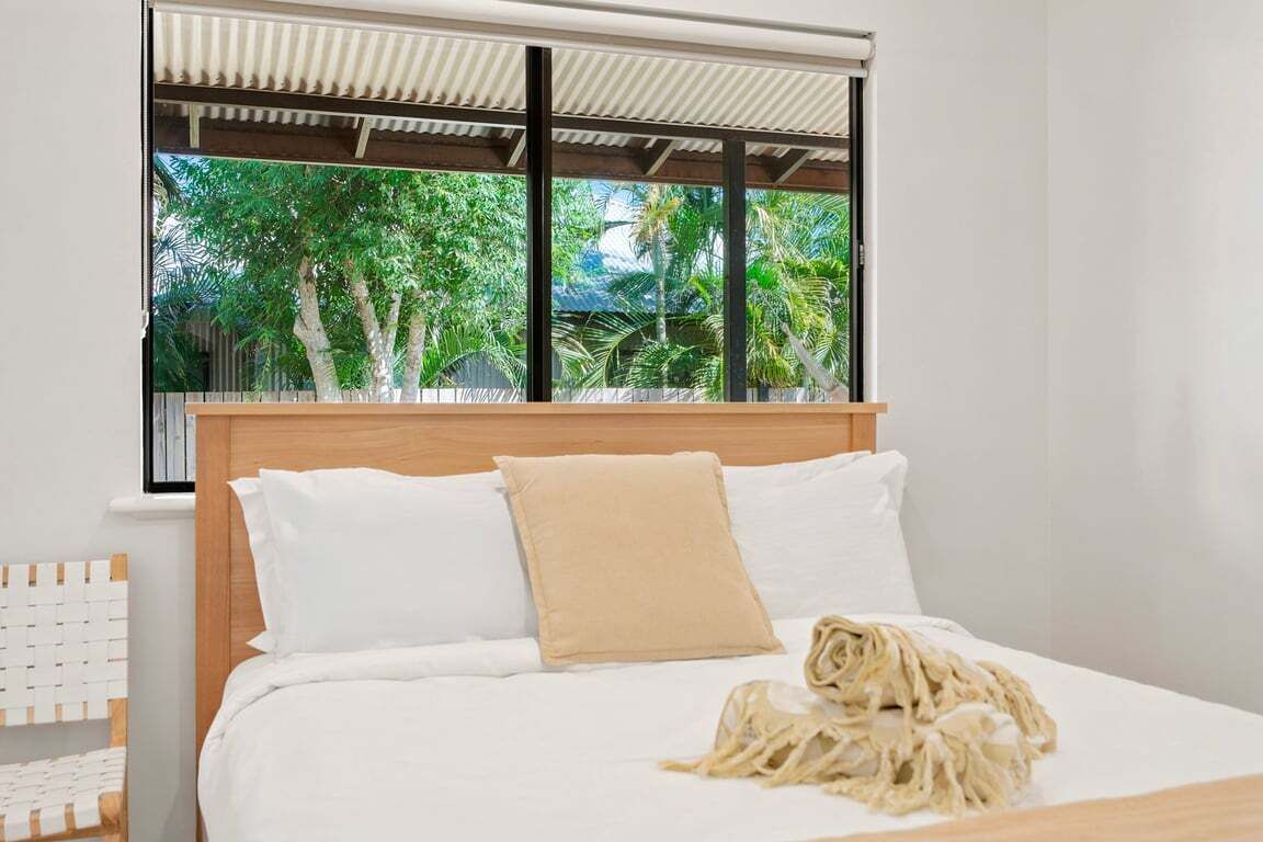 A bed with white bedding and a beige pillow, a wooden headboard, and a view of greenery through a window with a metal frame.